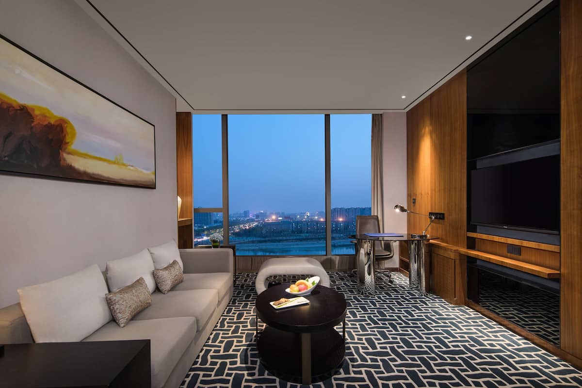 Een moderne hotelkamer met een groot raam met uitzicht over de stad, een bank, een ronde salontafel met fruit en een flatscreen-tv.