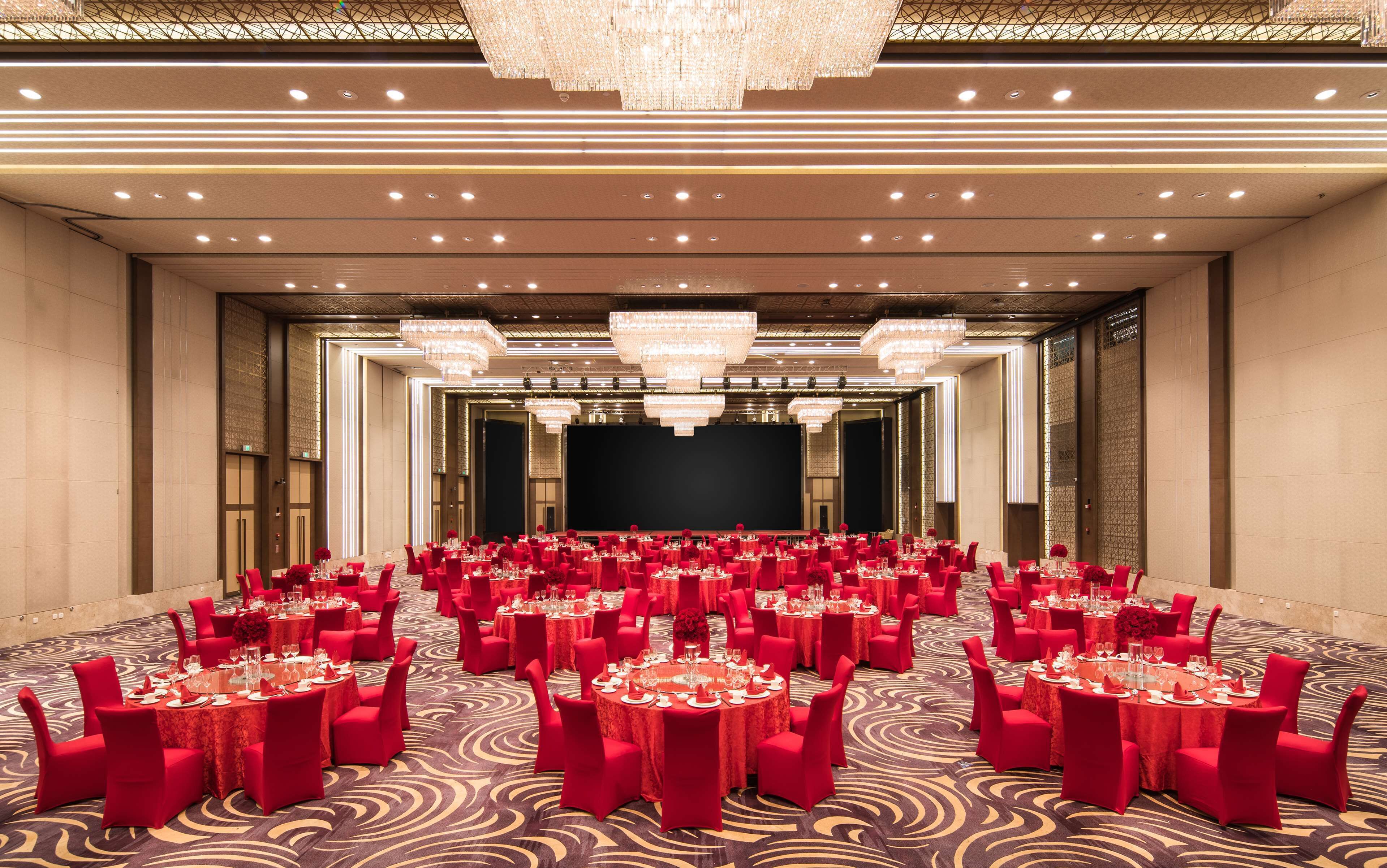 A banquet hall with round tables, red chairs, and chandeliers.