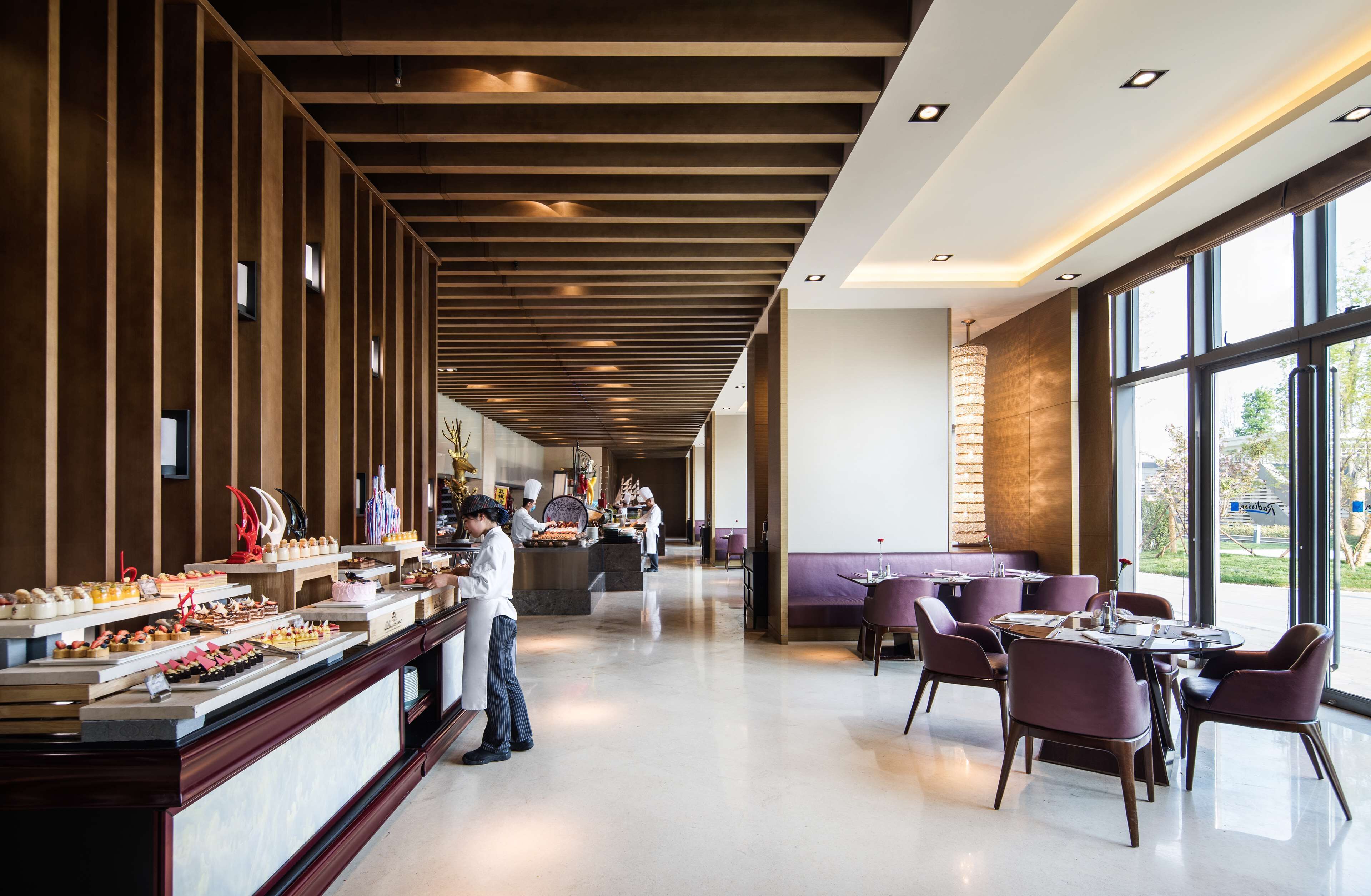 Un restaurant moderne doté d’un long comptoir où sont exposés différents plats, d’un espace avec des tables et des chaises, et de grandes fenêtres offrant une vue sur l’extérieur.