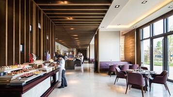 Restaurante moderno com um balcão extenso exibindo diversos pratos, área de estar com mesas e cadeiras, e grandes janelas oferecendo vista para o exterior.