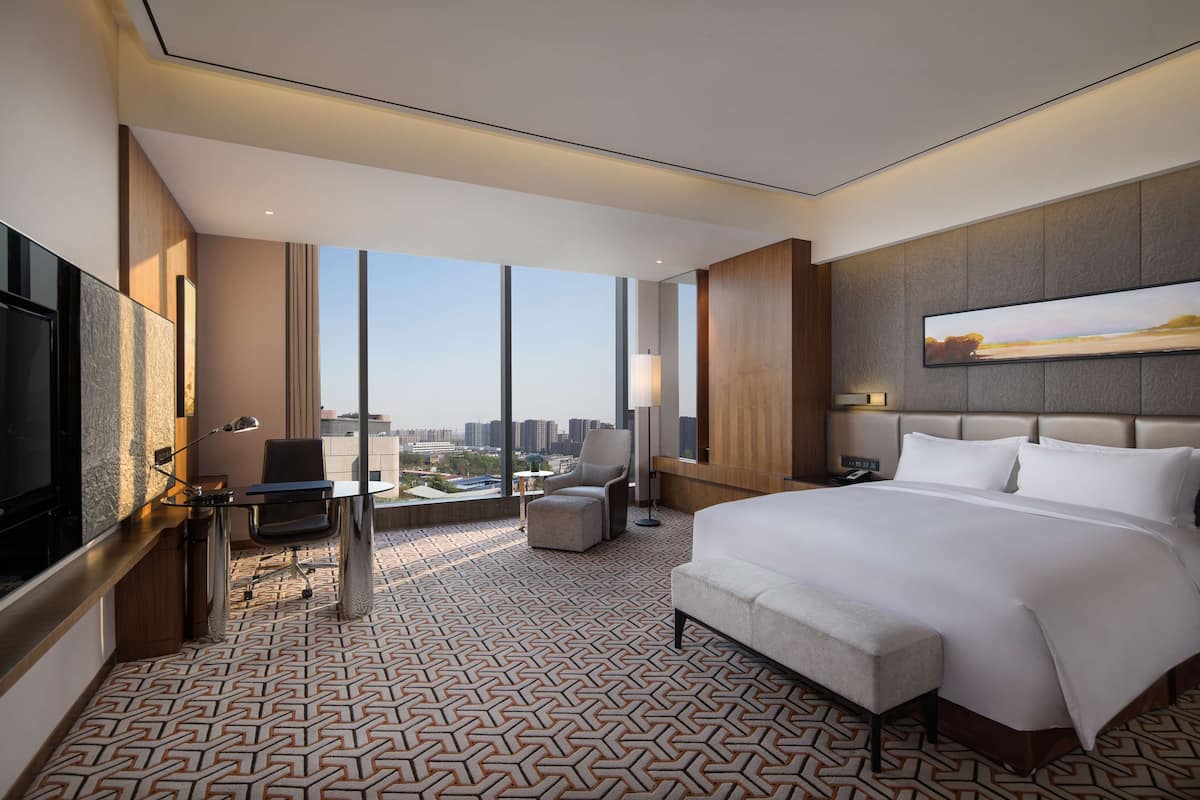 Hotelkamer met een groot bed, een bureau met stoel, een televisie en een groot raam met uitzicht op de stad.