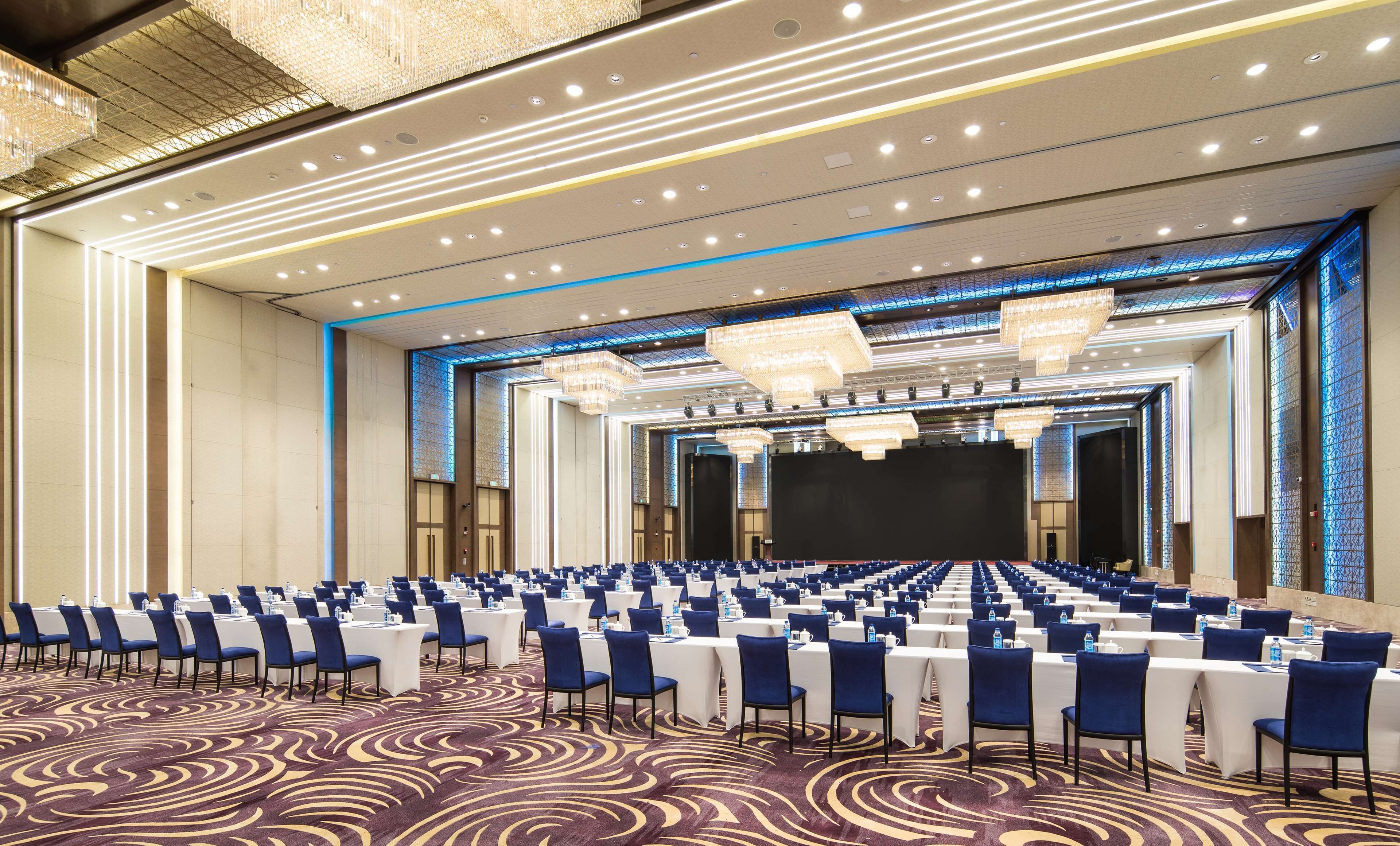 A large conference room with rows of tables and chairs, a stage, and a patterned carpet.