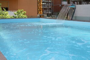 A pool with a waterfall feature, surrounded by a building with a brick facade and a metal structure in the background.