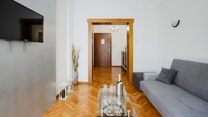 Ein modernes Wohnzimmer mit grauem Sofa, einem flachbildfernseher an der Wand, einer Holztür, die in einen anderen Raum führt, und einem Glastisch mit einer Flasche und Gläsern.