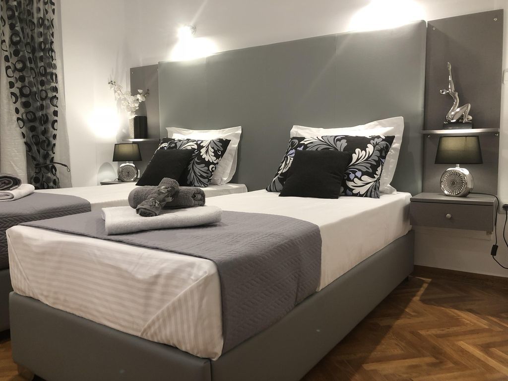 A modern hotel room with two beds, grey bedding, a wooden floor, and a nightstand with a lamp and decorative items.