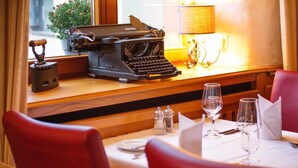 Un coin repas avec une machine à écrire ancienne posée sur une table en bois, des salières et poivrières, des verres à vin et des chaises à revêtement rouge.