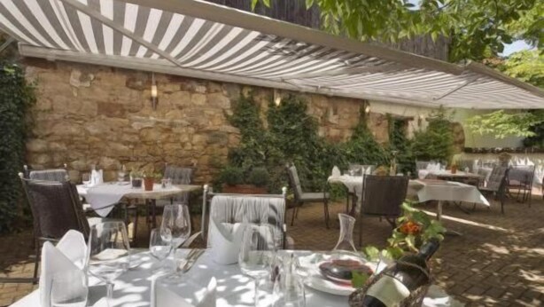 Un espace de restauration en plein air, avec des tables dressées pour le repas, protégé par un auvent à rayures et entouré de murs en pierre et de verdure.