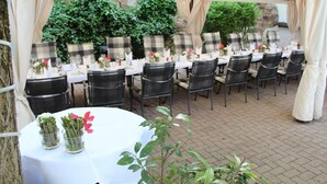 Un espace de restauration en plein air avec des tables rondes recouvertes de nappes blanches, des chaises en métal noir et des coussins à carreaux.