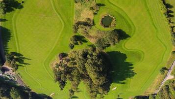 An aerial view of a golf course with a water body at the bottom, surrounded by trees and a road.