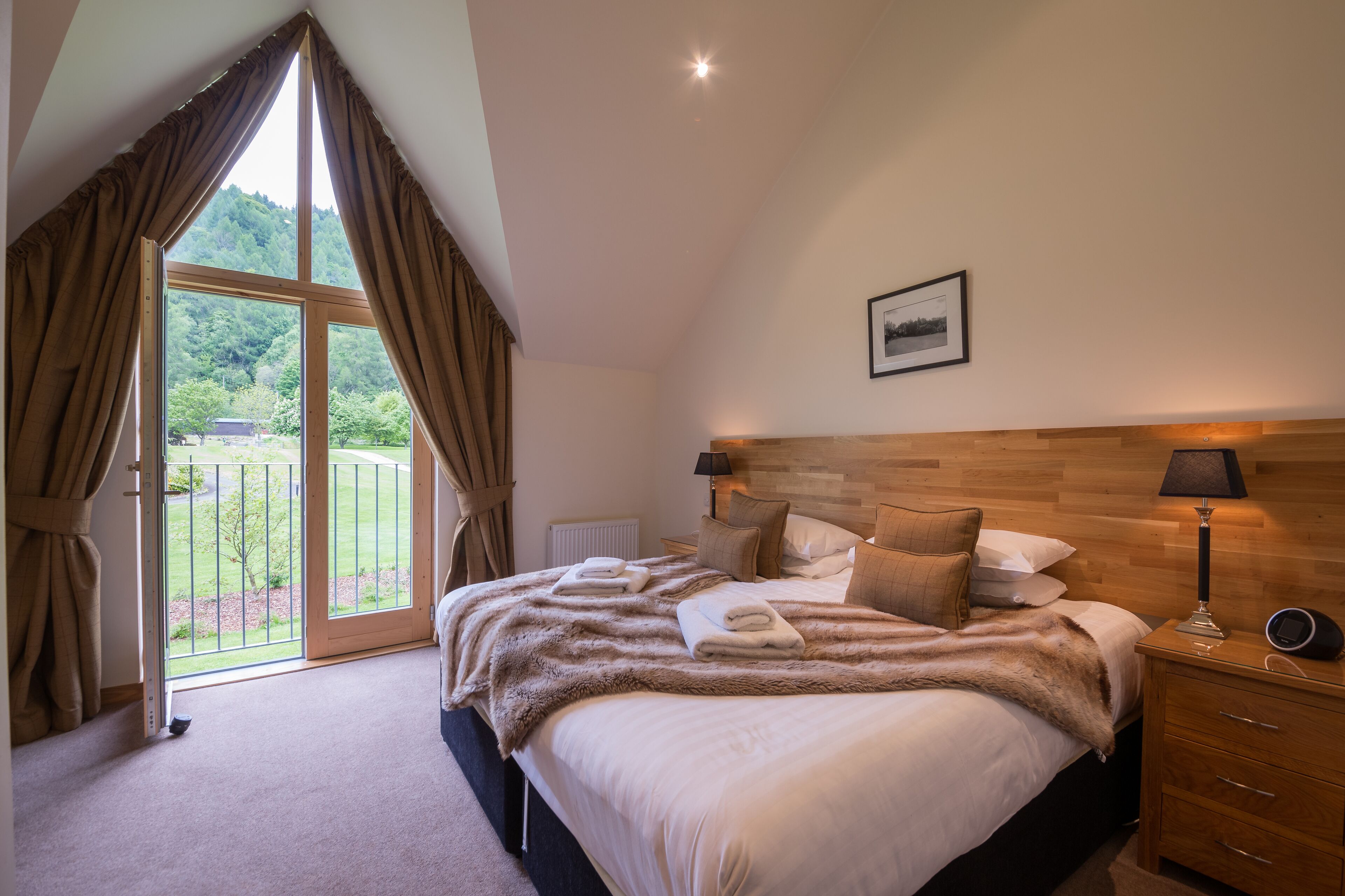 A bedroom with a large bed, a wooden headboard, a nightstand with a lamp, and a view of a garden through a sliding door.