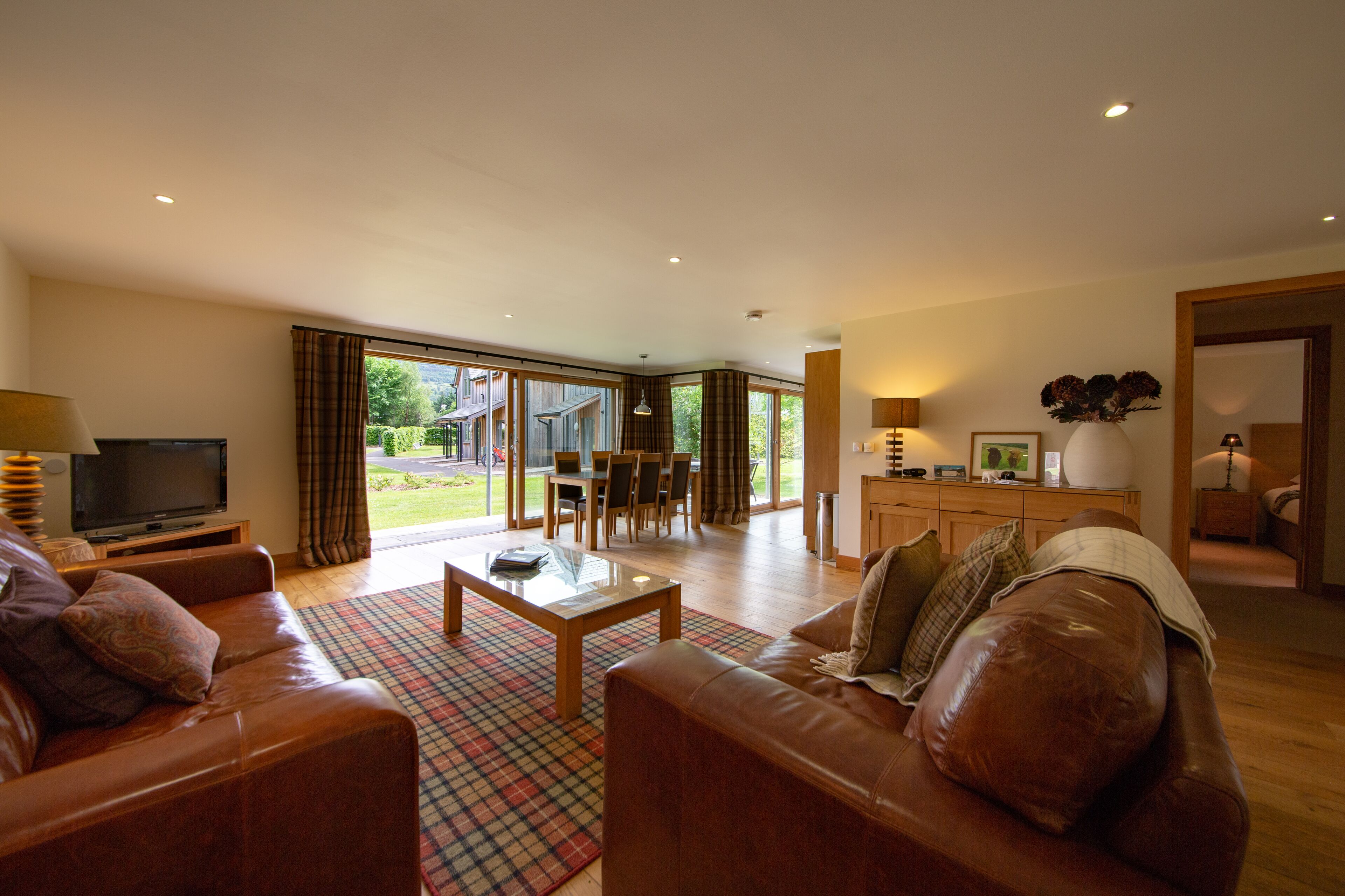 A spacious living room with leather sofas, a coffee table, and a TV. There is a dining area with a table and chairs, and large windows offering a view of the outdoors.