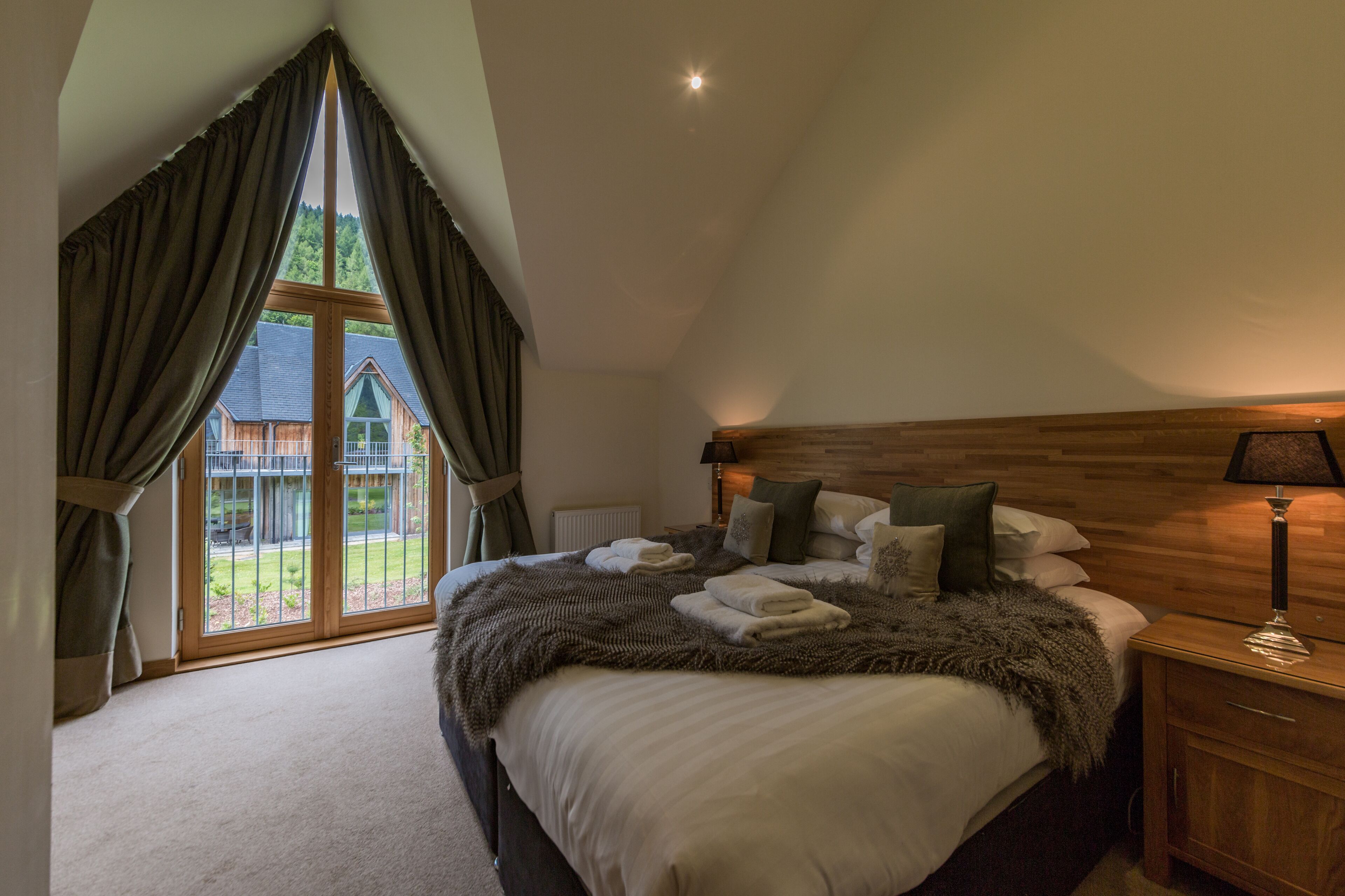 Un dormitorio con una cama grande, cabecera de madera y vistas a una cabaña de madera a través de una ventana con cortinas.