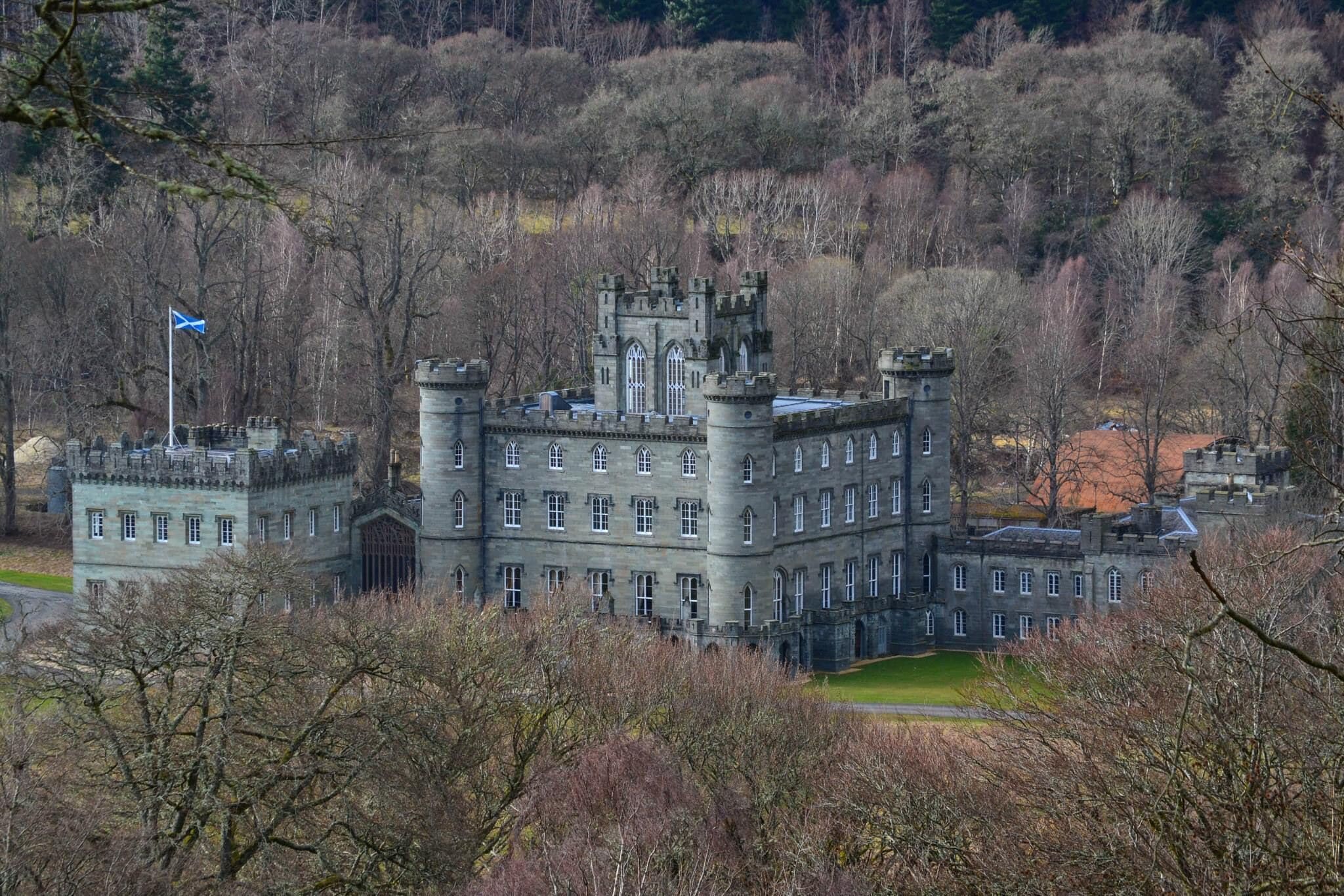 A large, historic stone building with multiple towers and arched windows, surrounded by trees and a flag.