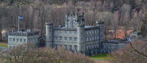 A large, historic stone building with multiple towers and arched windows, surrounded by trees and a flag.