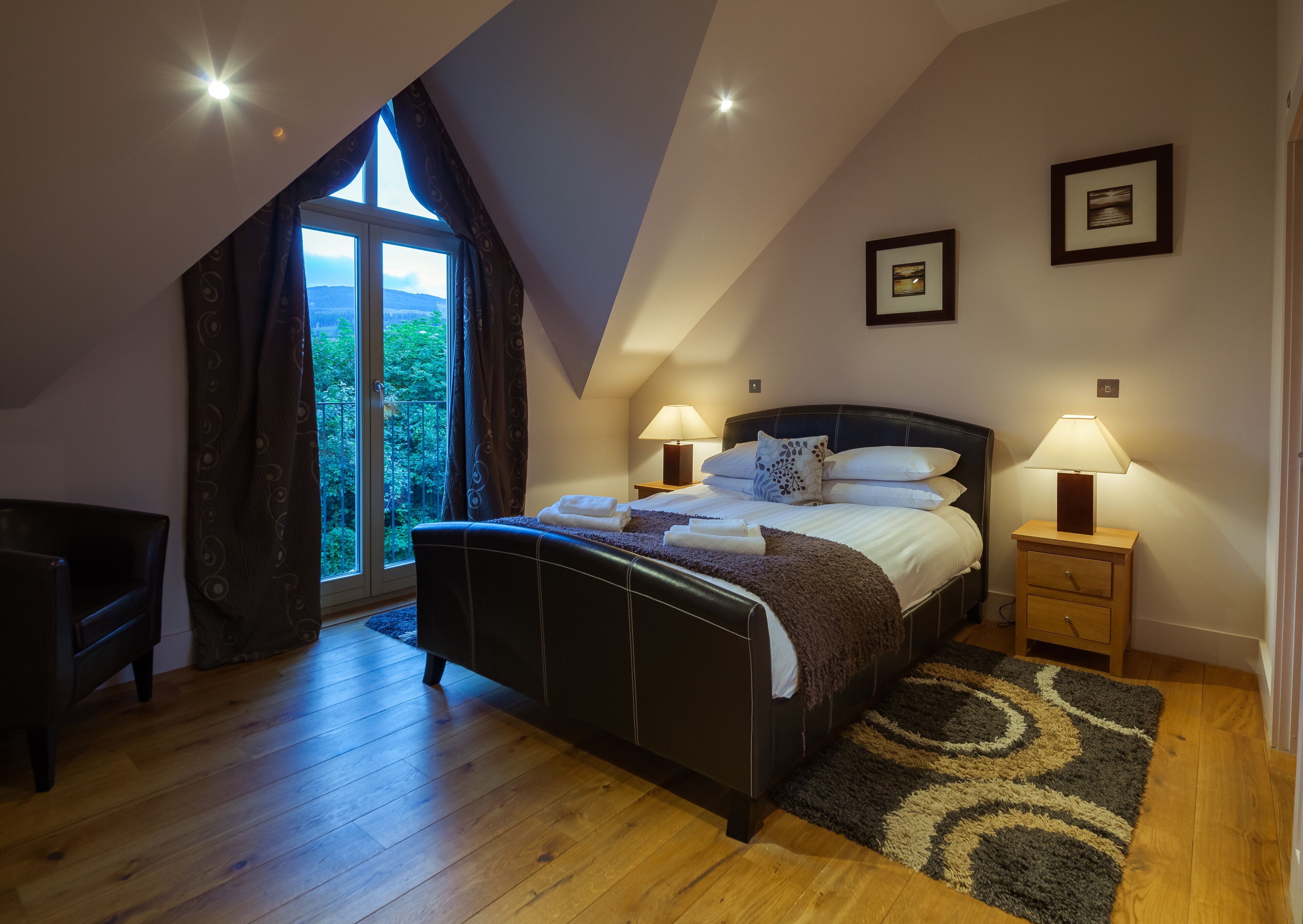 A bedroom with a large bed, two bedside lamps, a nightstand, a chair, and a view of greenery through a window.