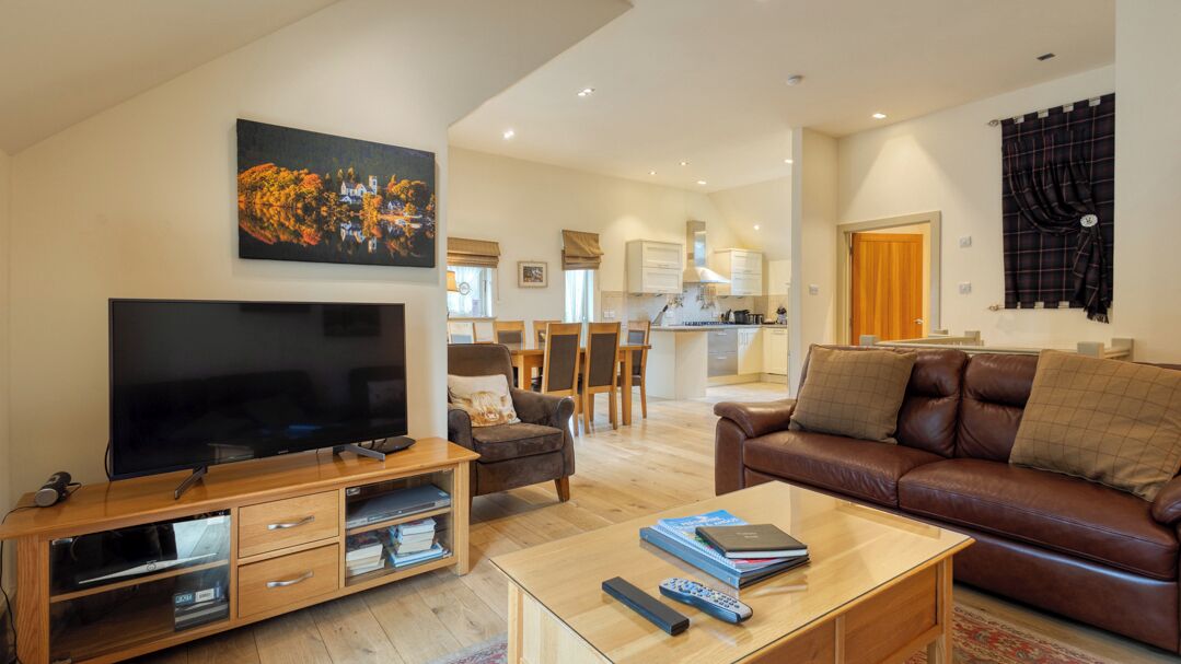 A modern living room with a flat-screen TV, a leather sofa, a coffee table with books, and a dining area in the background.