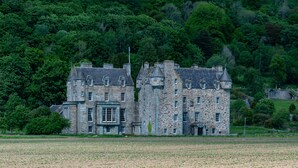 Un grand bâtiment en pierre, doté de nombreuses tours et d’une entrée centrale, entouré d’un champ et d’une forêt dense.