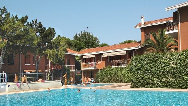 A swimming pool with people swimming and sunbathing, surrounded by buildings with balconies and a hedge.