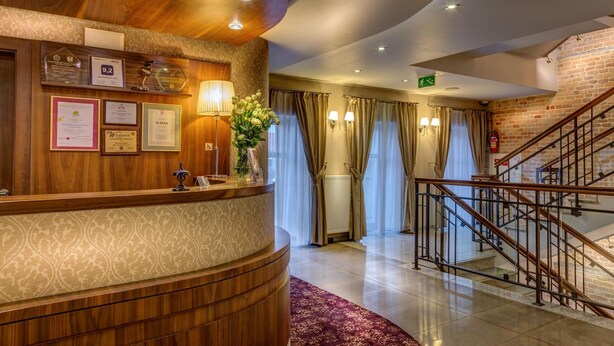 A hotel reception area with a wooden reception desk, framed certificates, a vase with flowers, a staircase, and a brick wall.