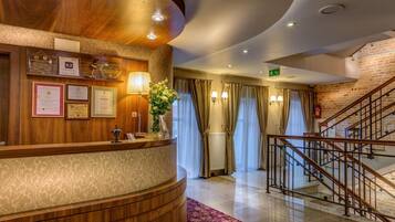 A hotel reception area with a wooden reception desk, framed certificates, a vase with flowers, a staircase, and a brick wall.