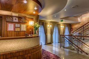 A hotel reception area with a wooden reception desk, framed certificates, a vase with flowers, a staircase, and a brick wall. - Hotel-restauracja Platan (Chrzanow)