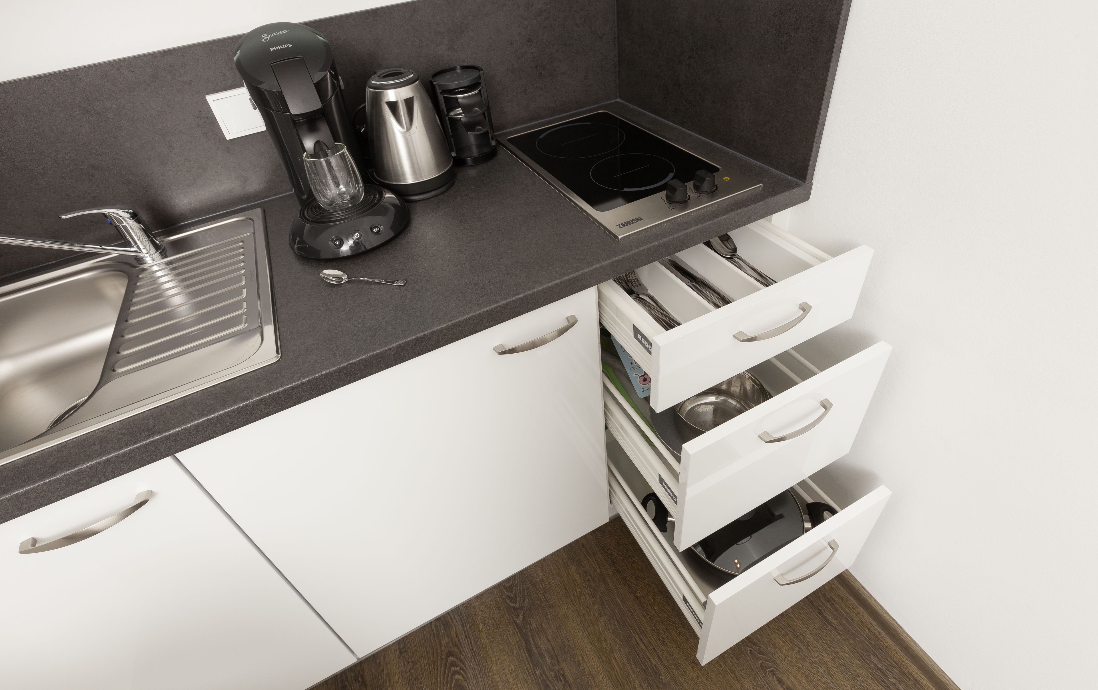 A modern kitchen with a built-in sink, a coffee maker, a kettle, and a stove. There are white cabinets and a drawer unit with multiple compartments.