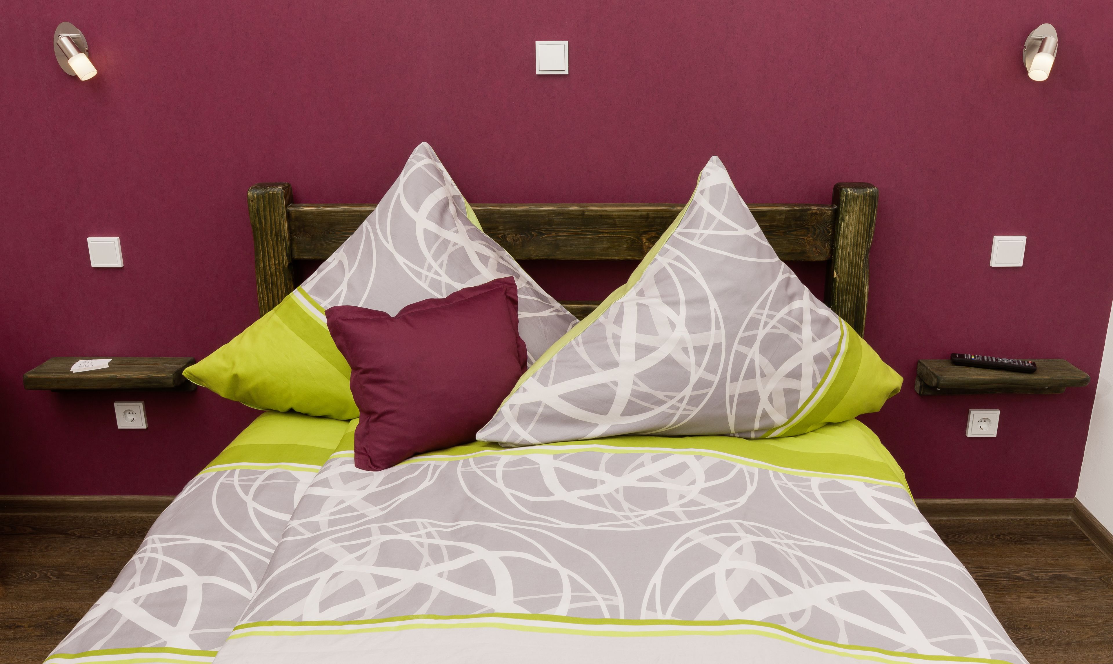 A bed with a patterned bedspread, two pillows, and a wooden headboard. There are bedside tables with remote controls and a wall socket.