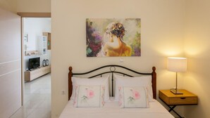 Une chambre à coucher avec un lit, des oreillers, une table de chevet avec une lampe, un tableau au mur et une vue sur la salle de bain et la cuisine.