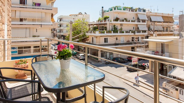 Un balcon avec une table et des chaises en verre, donnant sur une rue bordée d’immeubles et de voitures garées.