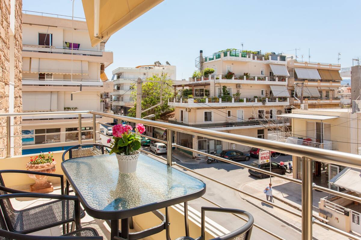 Un balcon avec une table et des chaises en verre, donnant sur une rue bordée d’immeubles et de voitures garées.