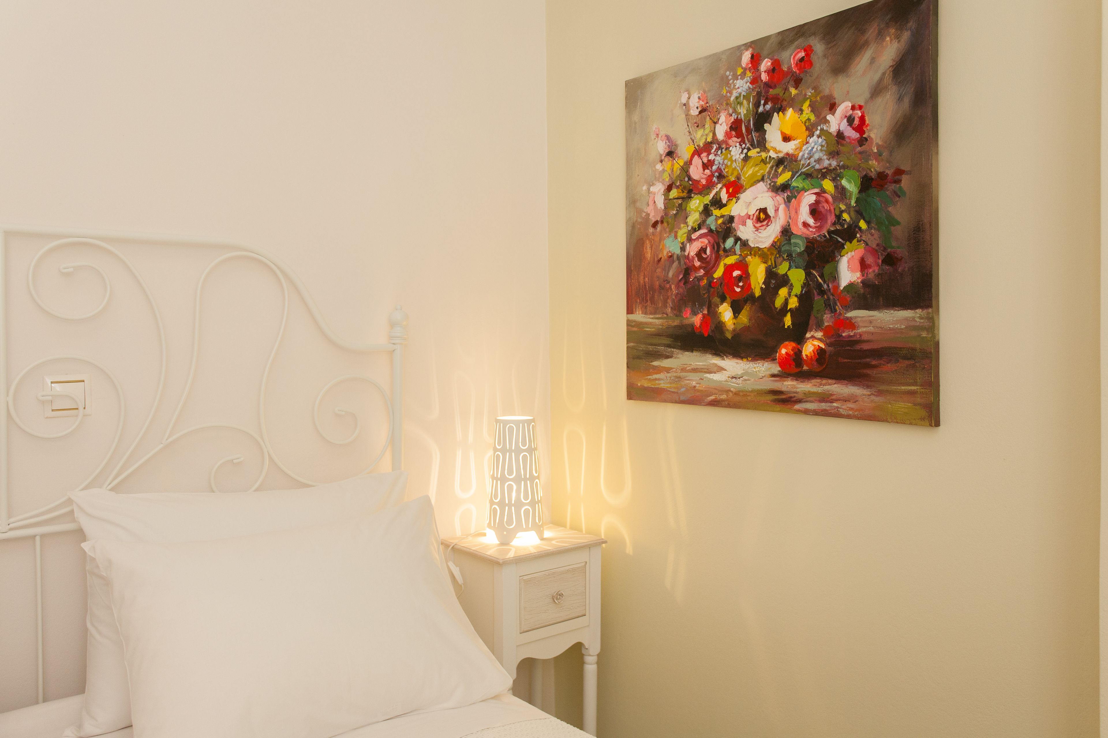 A bedroom with a white bed, a nightstand with a lamp, and a painting of flowers on the wall.