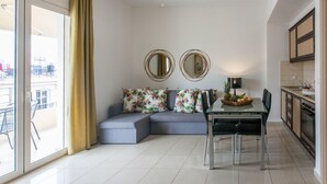 Un salon moderne avec un canapé d’angle gris, deux miroirs ronds, une table à manger avec des chaises et un coin cuisine équipé d’un évier et d’une cuisinière.