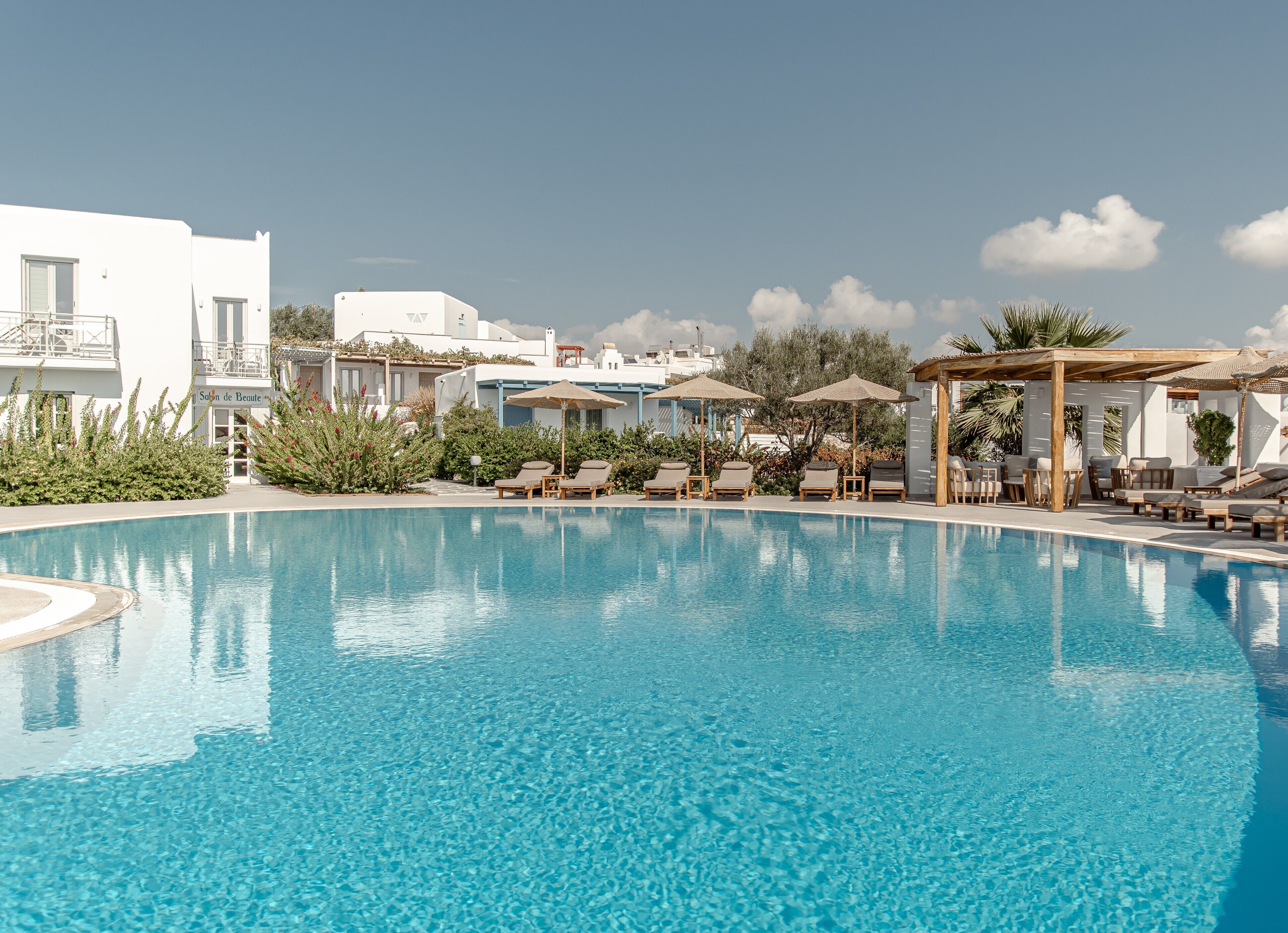 A clear blue swimming pool with lounge chairs and umbrellas, surrounded by white buildings and greenery.