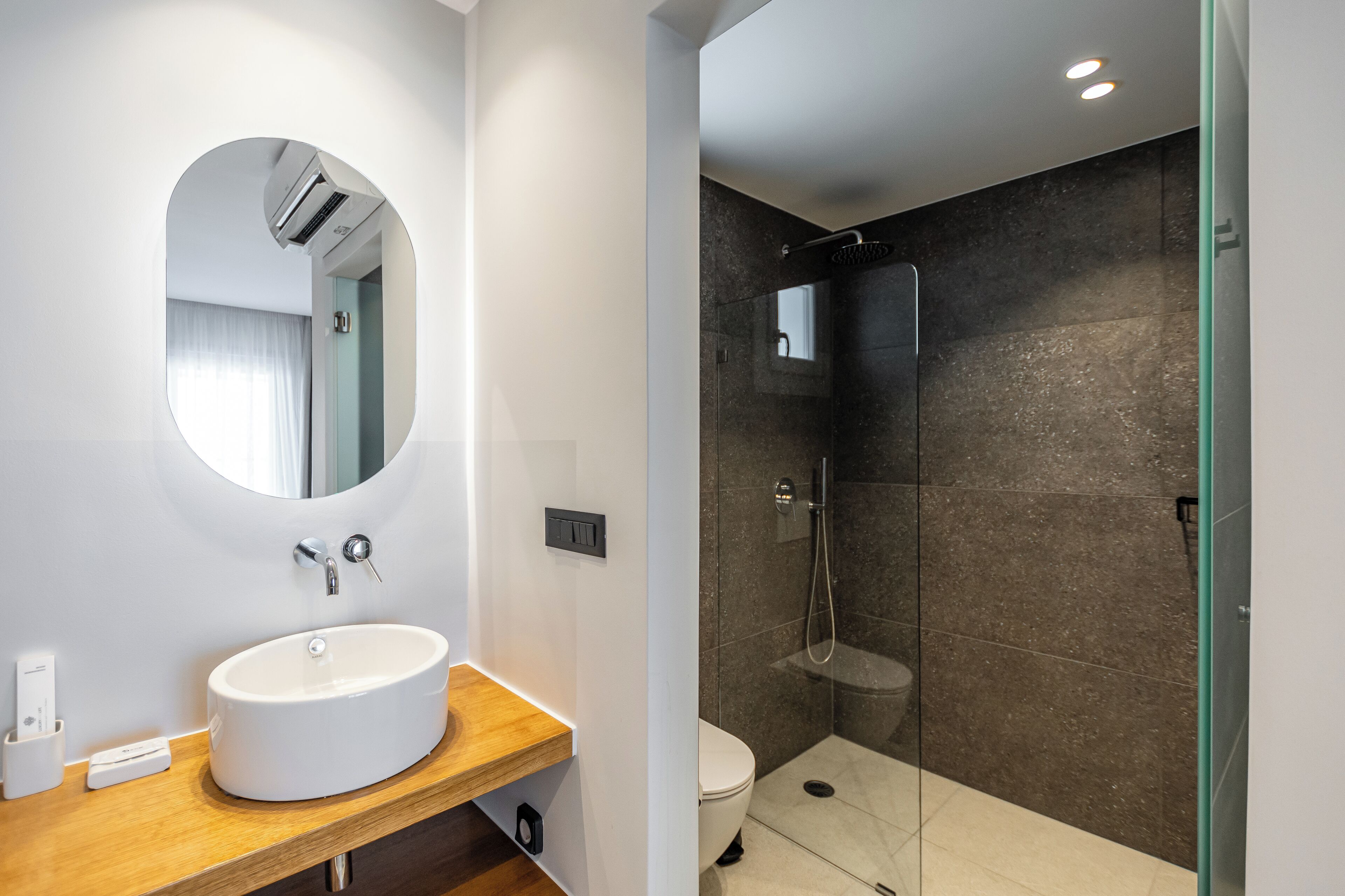 Un baño moderno con un espejo redondo, un tocador de madera, un lavabo blanco y una zona de ducha con mampara de vidrio.