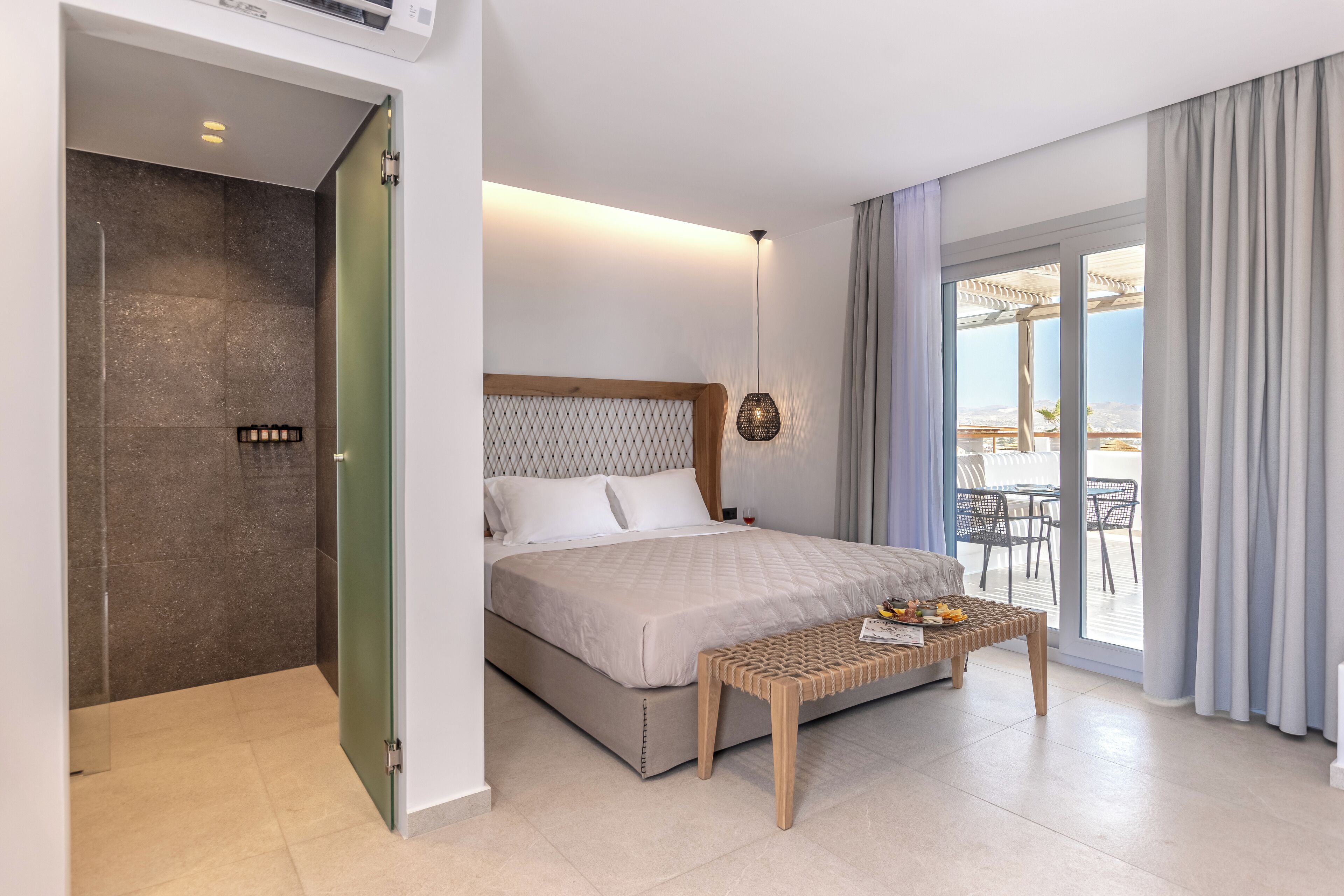 A modern hotel room with a bed, a small table, a shower area, and a balcony with a view.