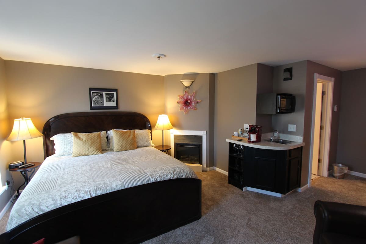 A bedroom with a large bed, bedside lamps, a fireplace, a kitchen area with a microwave, and a door leading to another room.