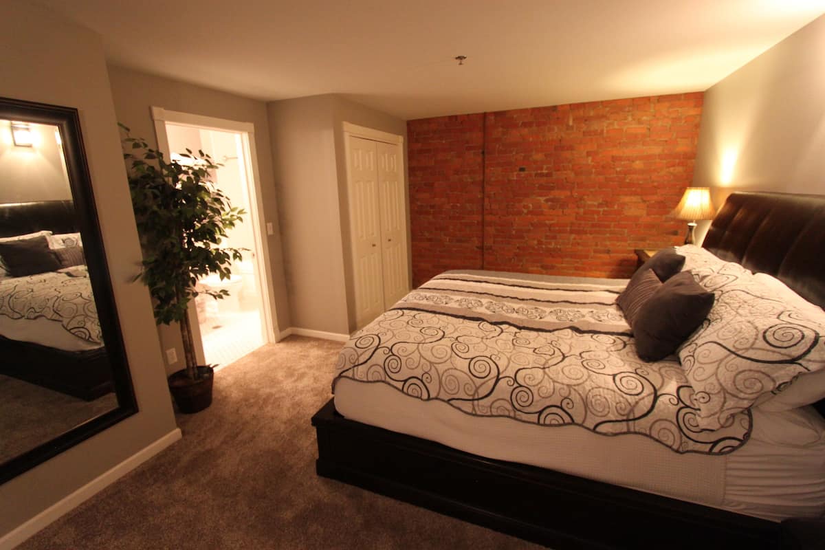 A bedroom with a large bed, a mirror, a potted plant, and a brick wall.