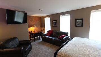 A room with a bed, a black leather sofa, a dining table with chairs, a TV mounted on the wall, and two framed pictures on the wall.