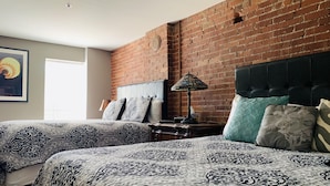 A bedroom with two beds, a brick wall, a headboard, a nightstand, a lamp, and a framed picture on the wall. - Lodging at the Gaffer Inn (Corning)