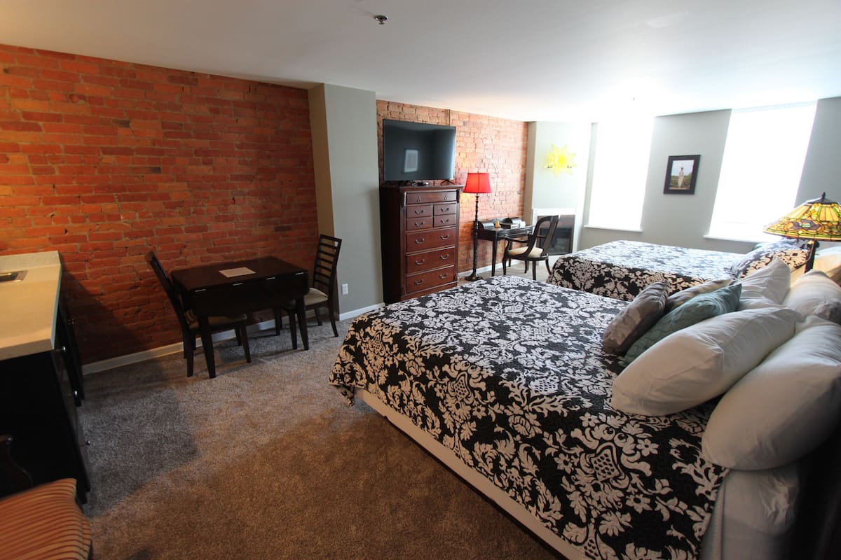 A room with a brick wall, a desk, a dresser, a bed with patterned bedding, and a chair.