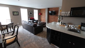 A living room with a sofa, a dining table with chairs, a kitchen area with a countertop and sink, and a TV mounted on the wall. - Lodging at the Gaffer Inn (Corning)