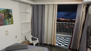 Une chambre d’hôtel avec un lit, une chaise, un balcon doté d’un sol à carreaux et une vue sur la ville la nuit.