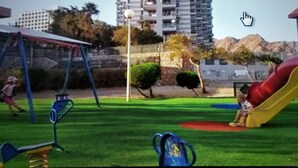 Une aire de jeux avec un toboggan et des balançoires, entourée d’une clôture et de verdure.