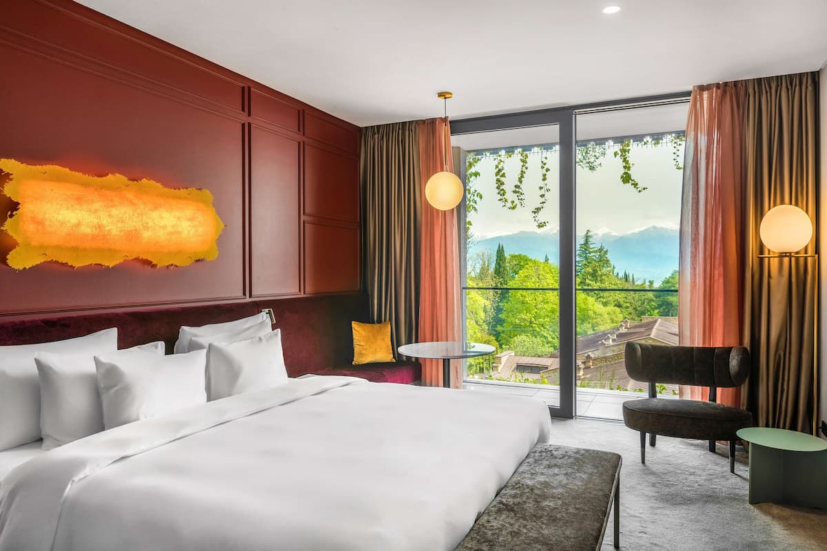 A hotel room with a large bed, a small table, a chair, and a view of a mountainous landscape through large windows.