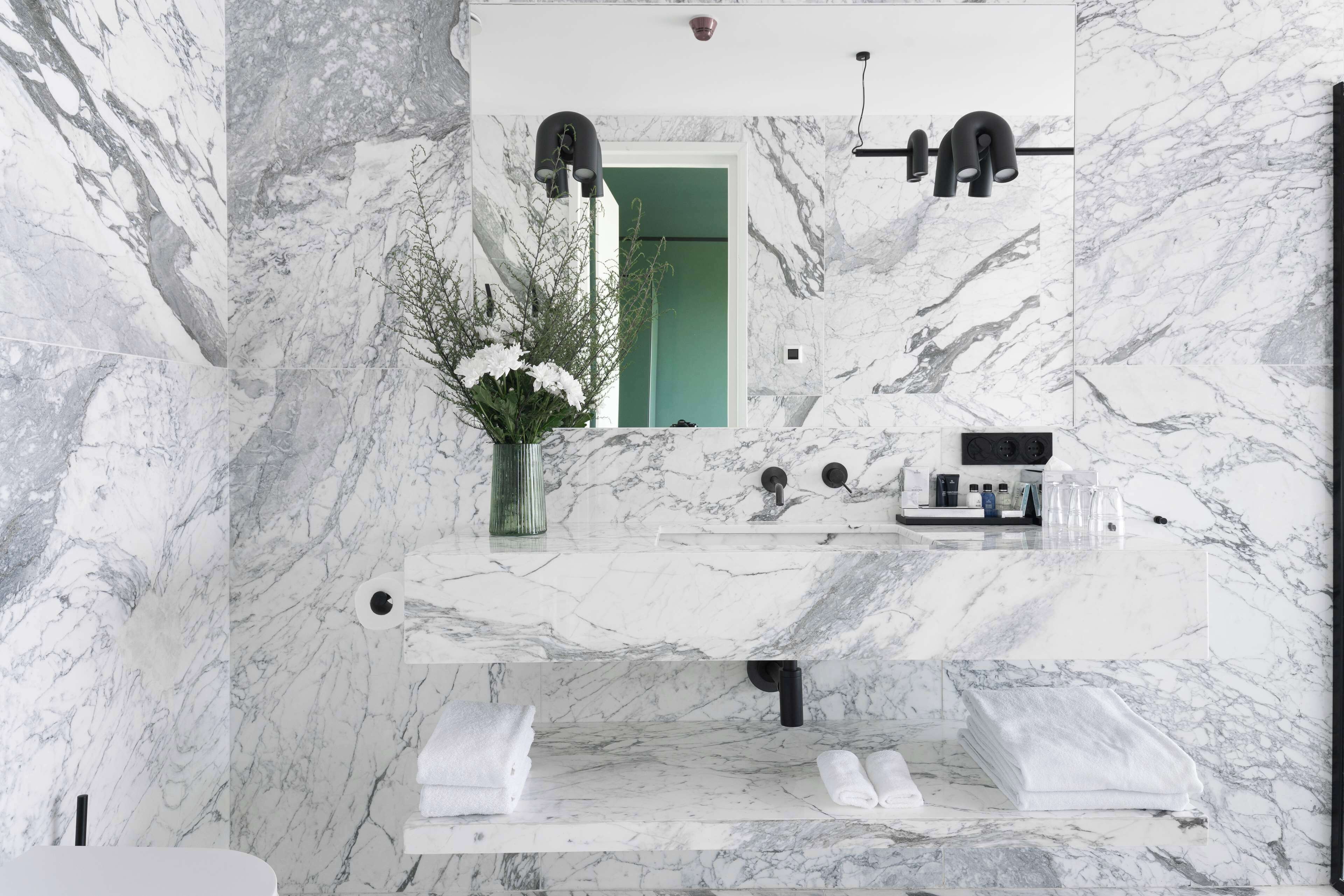 A bathroom with marble walls and a large mirror. There is a sink with a faucet, a vase with flowers, and a towel rack.