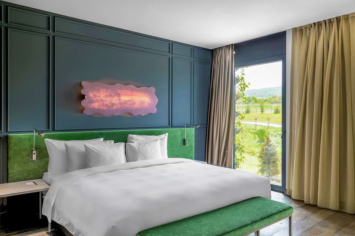 A hotel room with a large bed, a green upholstered bench, a wall-mounted light fixture, and a view of greenery through a window.
