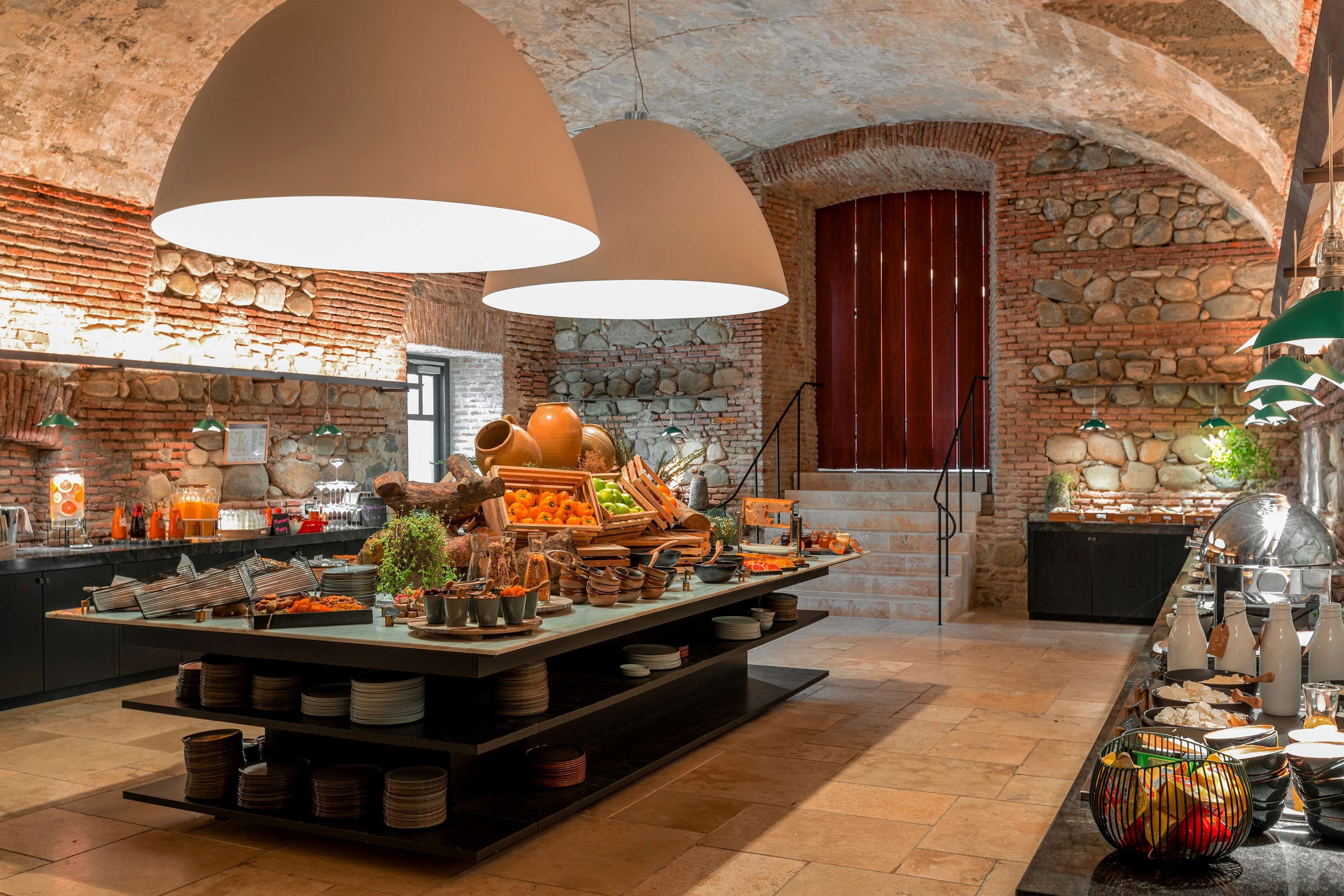 A breakfast buffet with a variety of food items and drinks, set in a rustic stone-walled room with arched ceilings and pendant lighting.