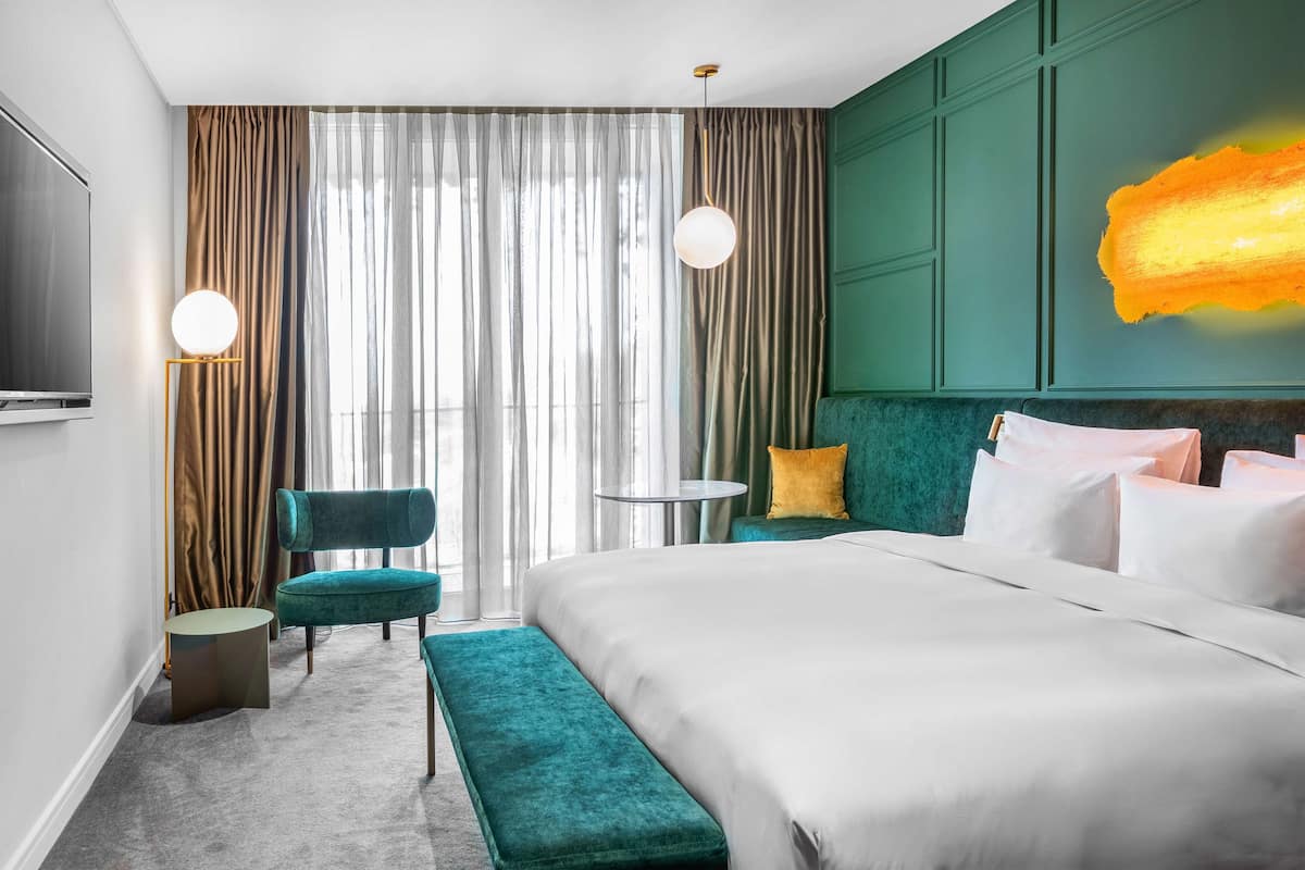A modern hotel room with a large bed, a green upholstered bench, a small round table, and a TV.