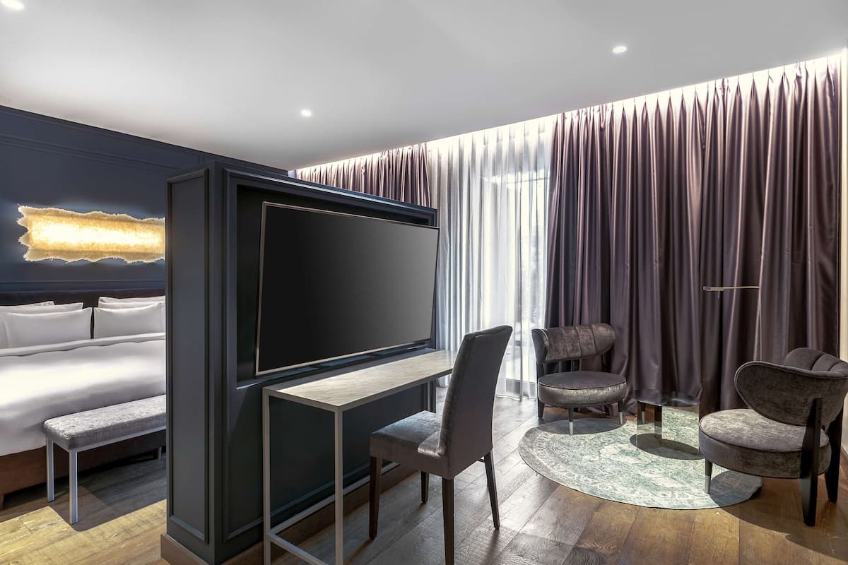A hotel room with a bed, a desk with a TV, two armchairs, a round rug, and a large window with curtains.