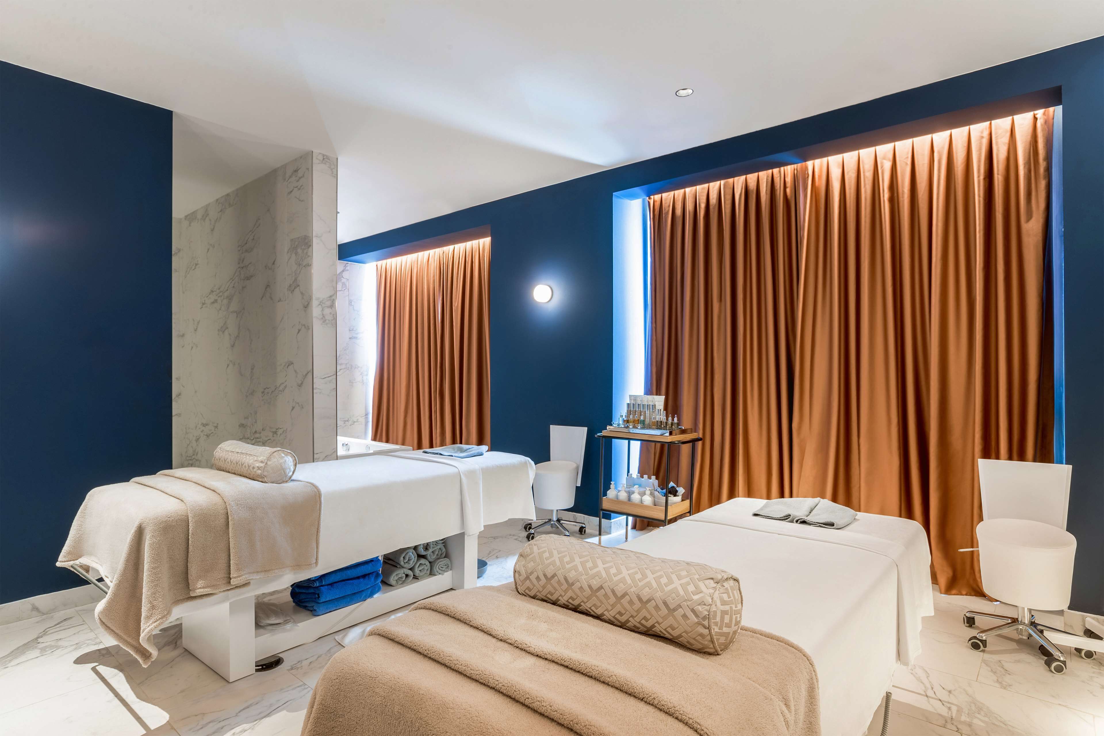 Una habitación de hotel moderna con dos camas, una mesa de masajes, una silla de masaje, una mesita con artículos de aseo y ventanas grandes con cortinas.